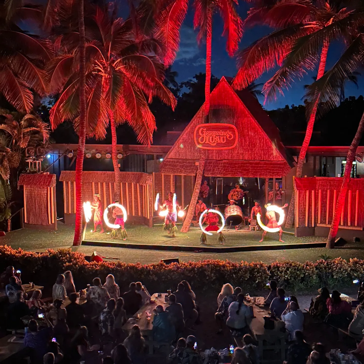 Night luau with fire dance and audience under palm trees.